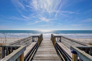Ein hölzerner Steg führt zu einem Strand mit blauem Himmel.