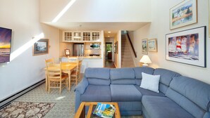 A modern living room with a blue sofa, wooden dining table, and chairs. There is a kitchen area in the background with a staircase leading to an upper level. - Beachfront 2 bedroom condo, Glen Arbor (Glen Arbor)