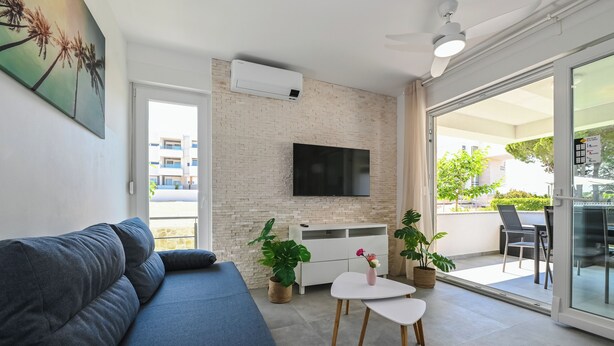 Una sala moderna con un sofá, un televisor de pantalla plana montado en una pared de ladrillo, una mesa pequeña con sillas y un balcón con mobiliario de exterior.