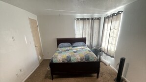 Une chambre à coucher avec un lit, un sol recouvert de moquette, un luminaire au plafond, une fenêtre avec des rideaux et une porte.