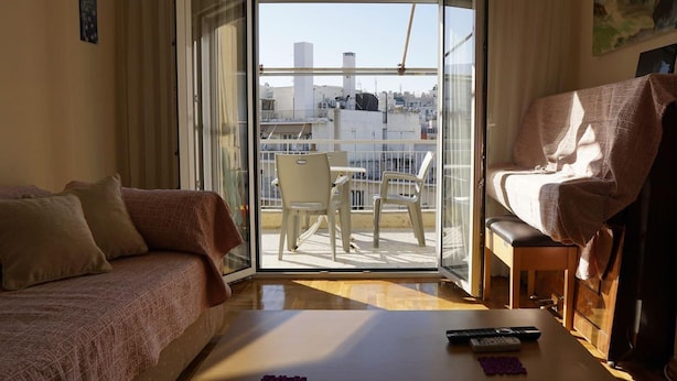 A hotel room with a bed, a sofa, a balcony with chairs, and a table with remotes. - Immaculate 2-bed Apartment in Zografou (Zografos)