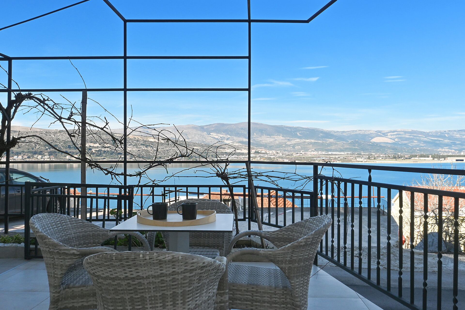 A terrace with wicker furniture, a table with two cups, and a view of a lake and mountains.