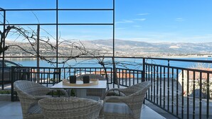 A terrace with wicker furniture, a table with two cups, and a view of a lake and mountains.