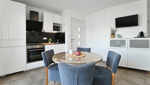 A modern kitchen with white cabinets, a built-in oven, and a microwave. There is a dining table with four chairs and a small potted plant on the counter.