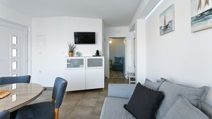 A modern living room with a grey sofa, a dining table with chairs, a TV mounted on the wall, and a kitchen area with cabinets and a plant.