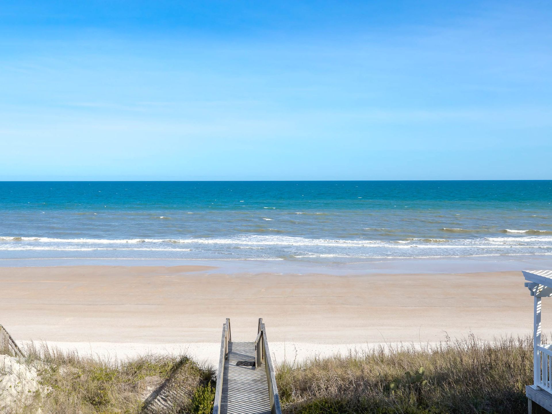 A beach with a wooden boardwalk leading to the shore, clear skies, and calm ocean waves.