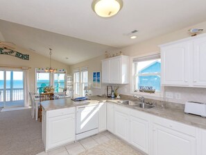 A kitchen with white cabinets, a central island, a view of the ocean through the windows, and a 'Welcome' sign on the window. - Attitude Adjustment  - 4BR Oceanfront Home; Great Views from the Covered Porches (Surf City)