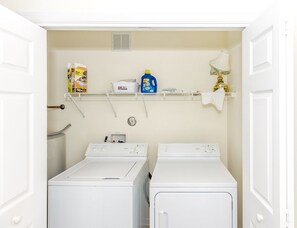 Une buanderie équipée de deux machines à laver et de deux sèche-linge à chargement frontal, tous deux de couleur blanche, d’une étagère contenant des produits de nettoyage et d’une serviette accrochée au mur.