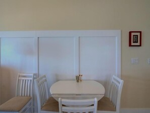A dining area with a white table and chairs, a small vase on the table, and a framed picture on the wall.