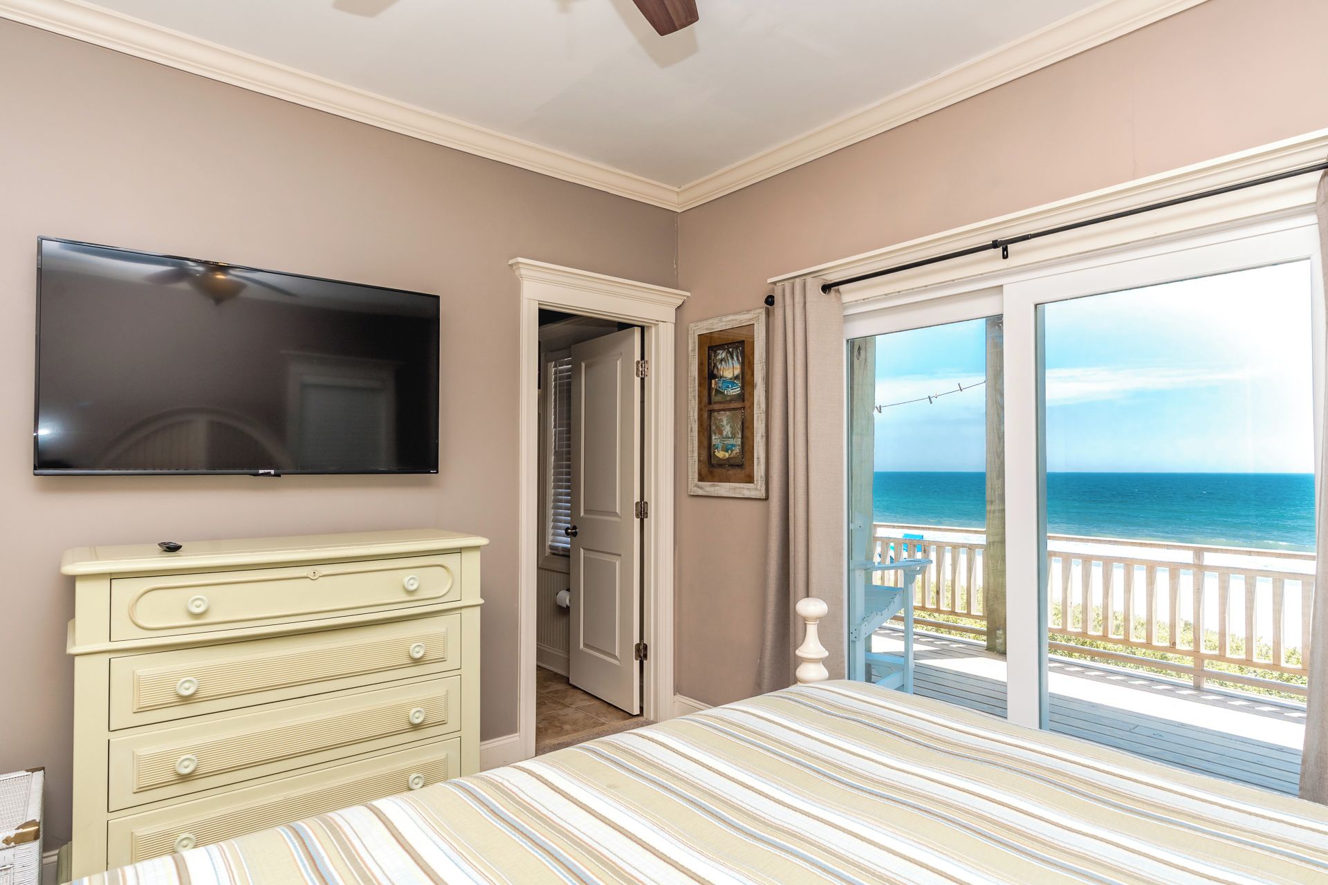 Un dormitorio con cama, tocador, televisor y puerta que da a un balcón con vistas al océano.