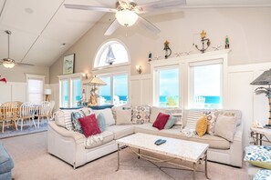 A spacious living room with a high ceiling, a ceiling fan, a large window with a view of the ocean, a dining area with a table and chairs, and a cozy seating area with a sofa and a coffee table.