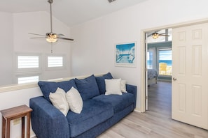 A room with a blue sofa, a ceiling fan, a framed picture, and a door leading to another room. - Que Sea-Ra Sea-Ra - 6BR Oceanfront Home. Private Pool & Hot Tub! Pet Friendly! (North Topsail Beach)
