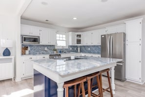 A modern kitchen with white cabinetry, a marble countertop, stainless steel appliances, and wooden stools. - Que Sea-Ra Sea-Ra - 6BR Oceanfront Home. Private Pool & Hot Tub! Pet Friendly! (North Topsail Beach)