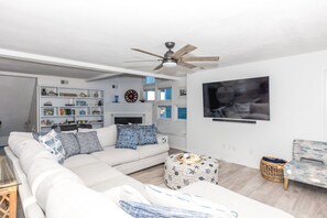 Un salon avec un canapé blanc, des coussins à motifs, une table basse, un téléviseur, une bibliothèque et un ventilateur de plafond.