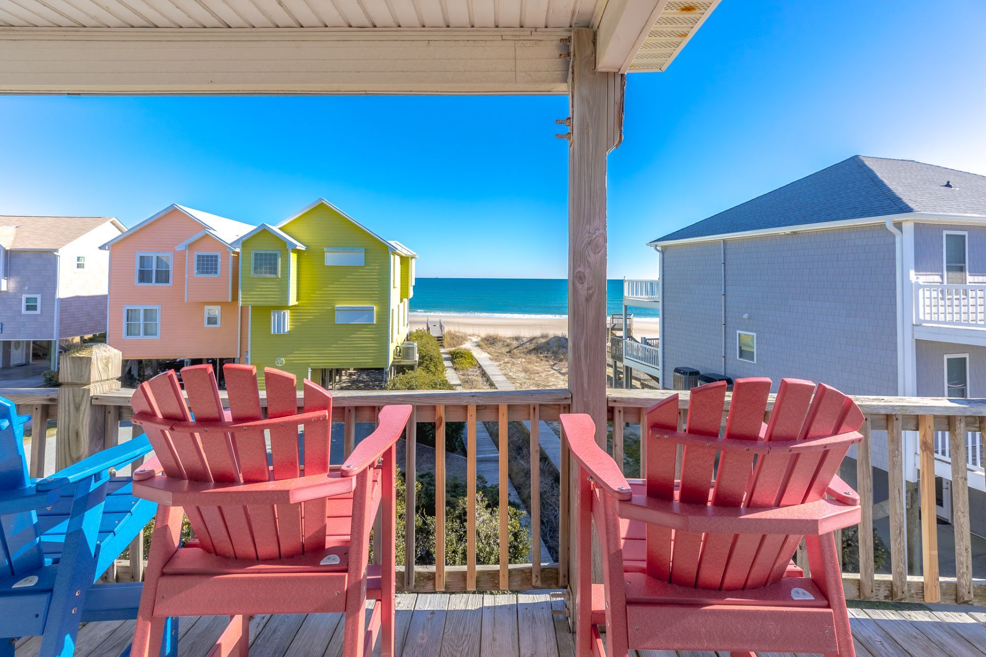 A wooden deck with red Adirondack chairs overlooking a beach with colorful houses.