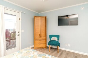Une pièce avec une armoire en bois, un fauteuil de couleur turquoise, un téléviseur fixé au mur et une vue sur l’extérieur par une porte.