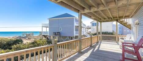 Une terrasse en bois offrant une vue sur la plage et l’océan, avec des rampes en bois et un espace abrité équipé de chaises.