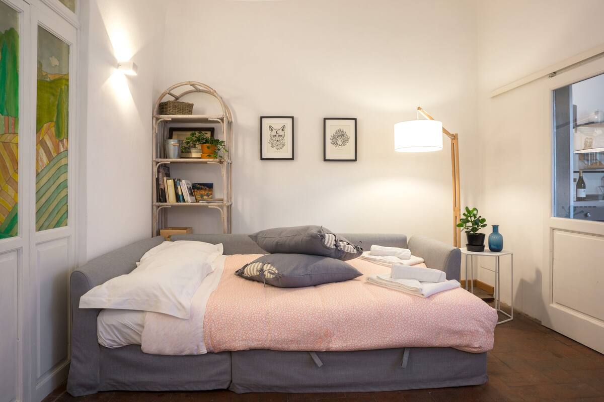 Een keurig opgemaakt slaapkamer met een bed, een nachtkastje met een lamp, een klein plankje met boeken en een plant, en lijstwerk aan de muur.