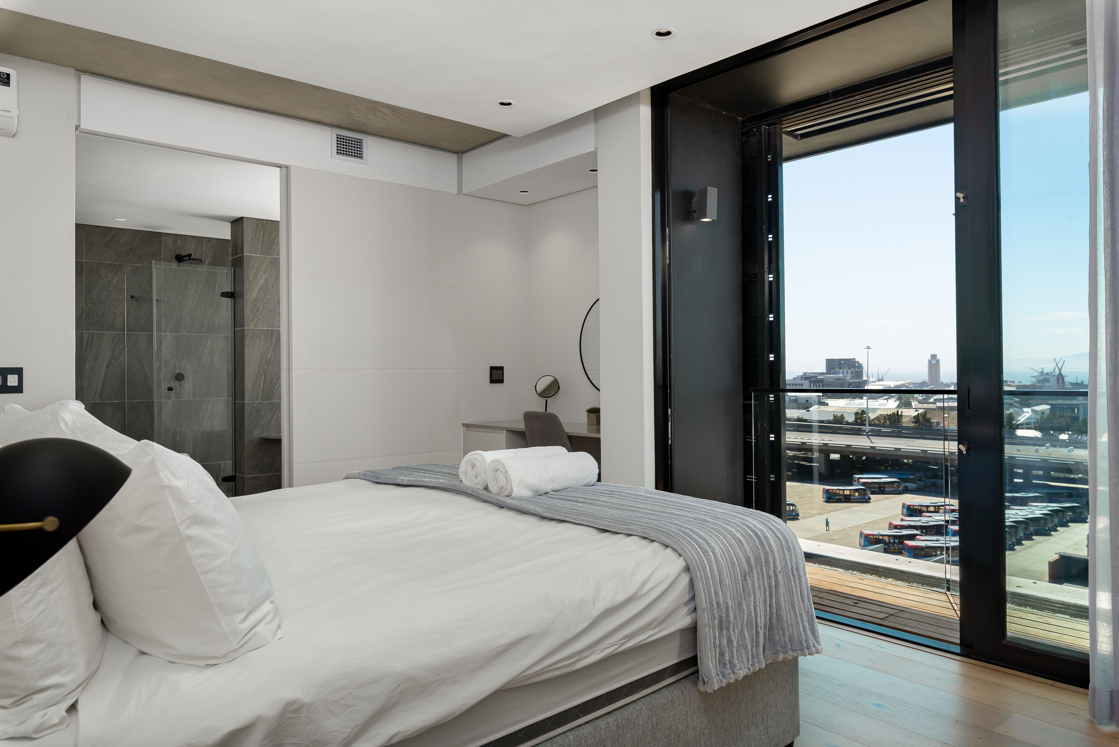 A modern hotel room with a large bed, a walk-in shower, a view of the city through a large window, and a desk with a chair.