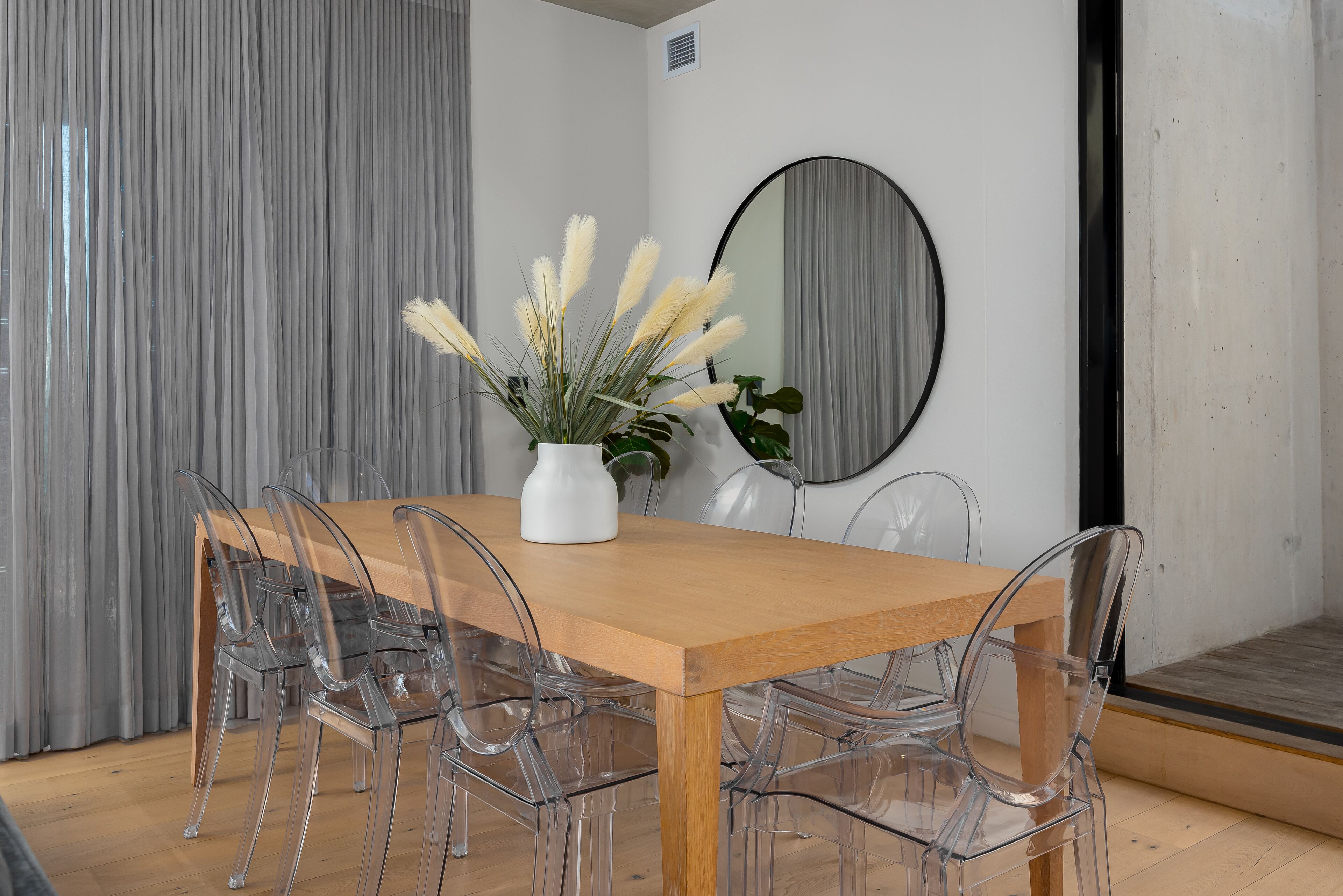 A dining area with a wooden table and clear plastic chairs, a white vase with pampas grass, and a round mirror on the wall.