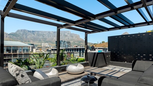 Un salón en la azotea con muebles modernos, una pérgola y vistas a edificios y montañas.