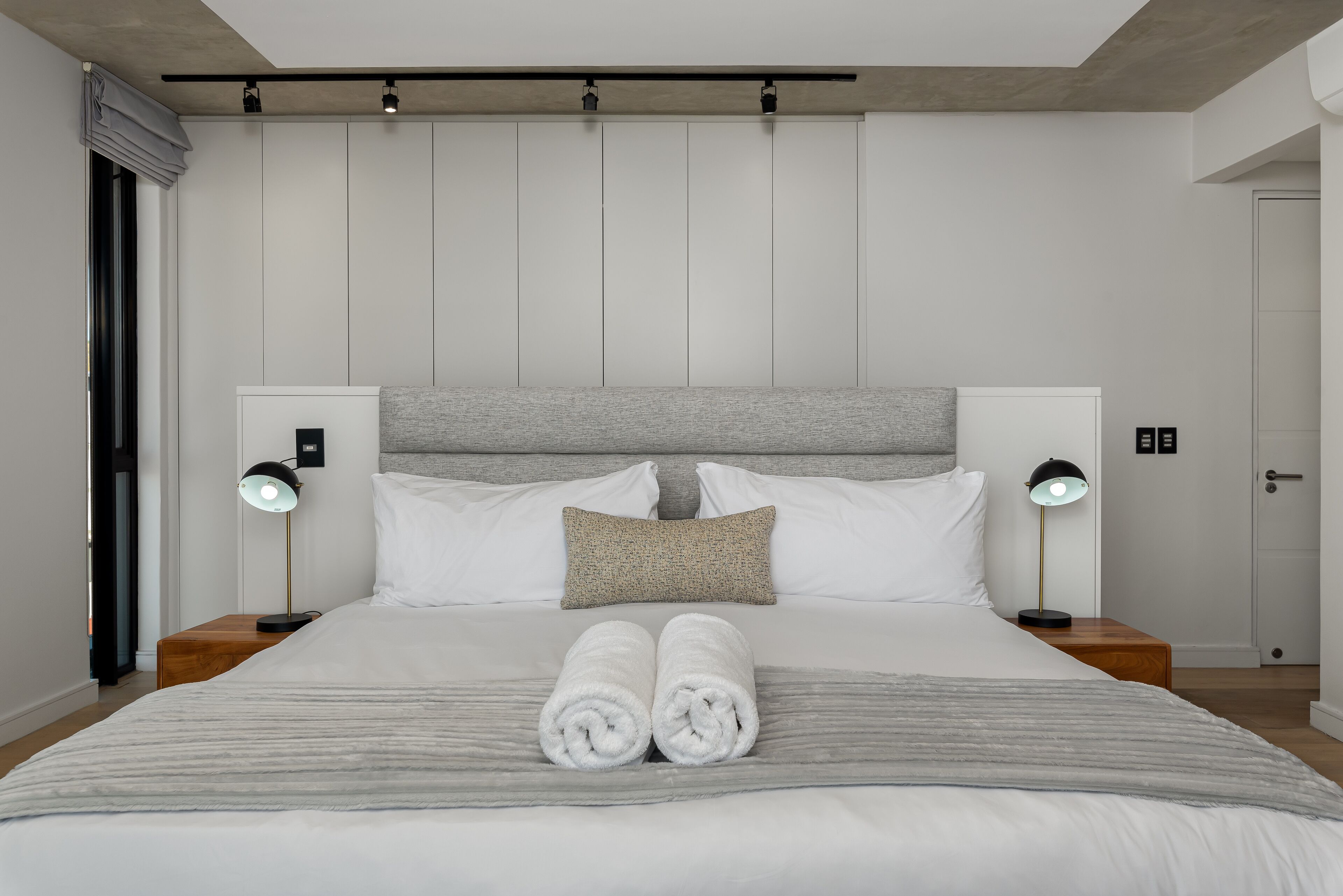 A modern hotel room with a large bed, two bedside tables with lamps, a headboard, and a door.