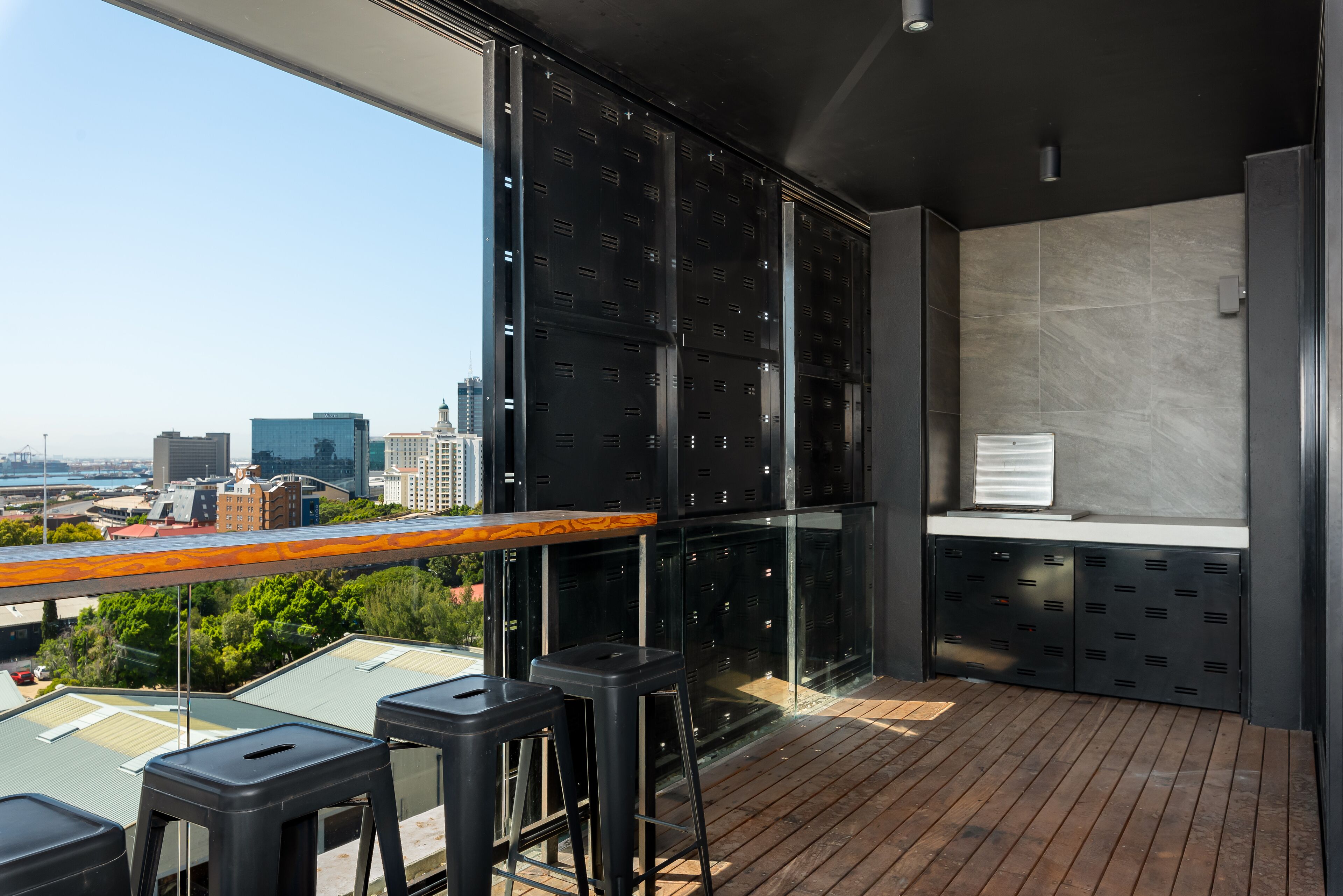 A modern bar area with a city view, featuring a wooden bar counter, black stools, and a large window with metal lockers.