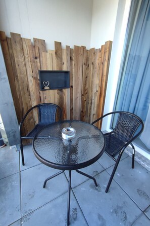Un petit espace extérieur aménagé avec une table ronde en verre et deux chaises en métal, une clôture en bois et un tableau noir orné d’un symbole en forme de cœur.