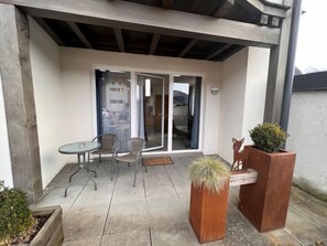 Une terrasse couverte avec une table et des chaises, un jardinier en bois contenant un arbuste et une sculpture de cerf.