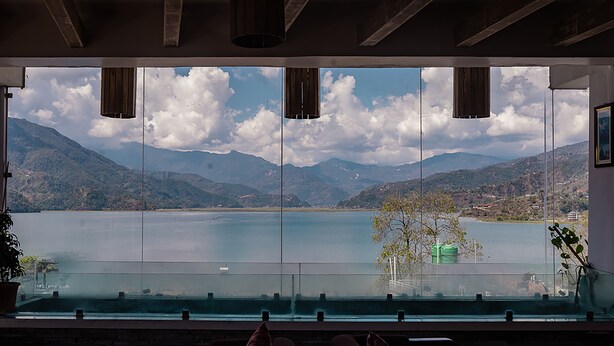 A room with a large window overlooking a lake and mountains. There are two hanging light fixtures and a potted plant on the windowsill. - Himalayan Glory Inn (Pokhara)