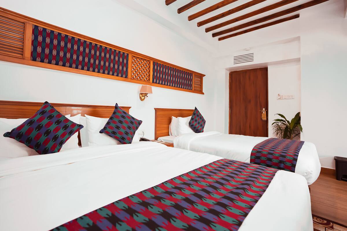 A hotel room with two beds, wooden headboards, patterned wall decor, a wall-mounted lamp, and a door with a handle.