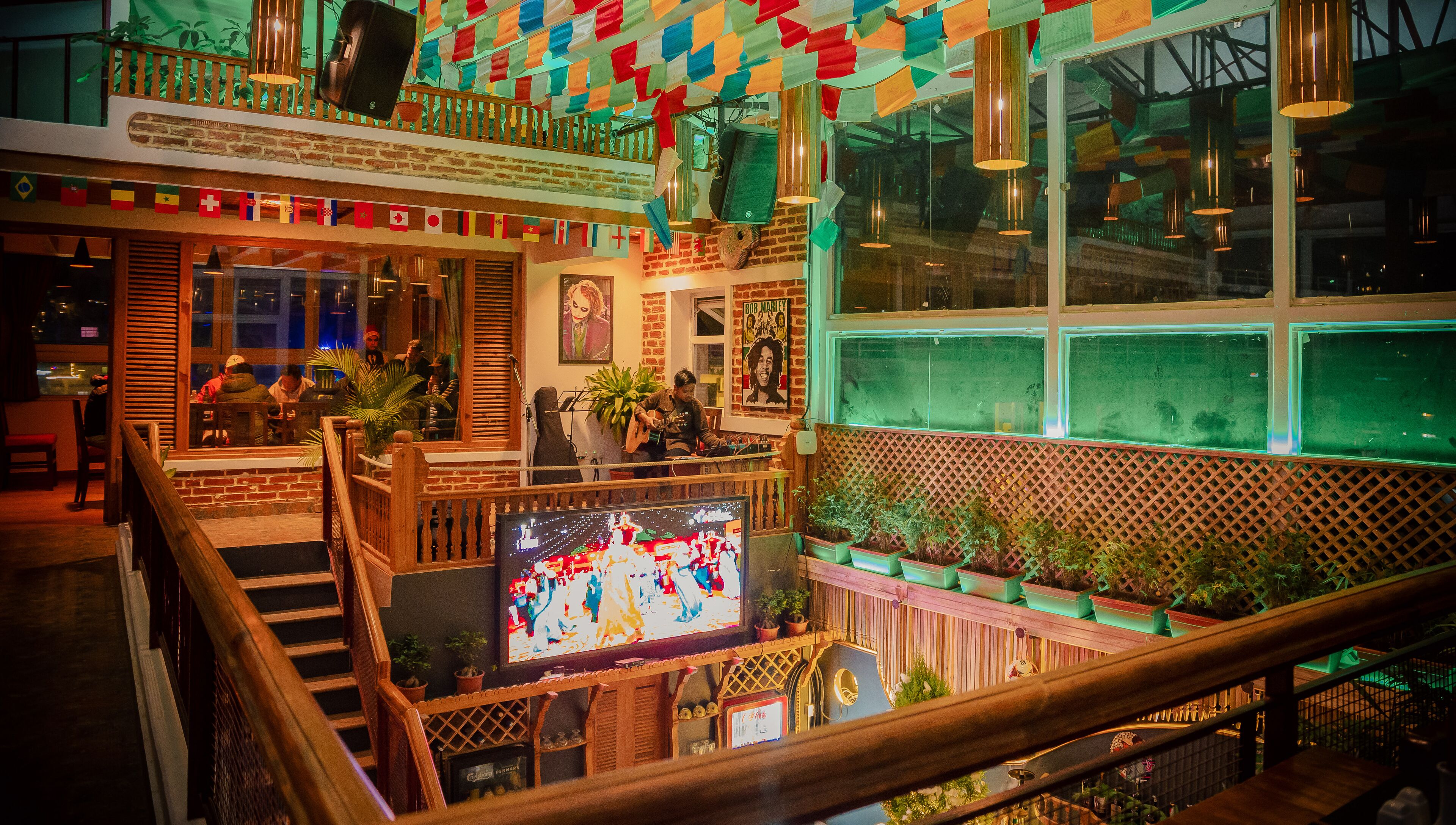 A restaurant with a wooden staircase, a TV screen displaying a show, and a balcony with colorful flags.