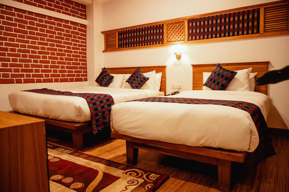 A hotel room with two beds, a brick wall, a wooden headboard, a patterned rug, and a wall-mounted light fixture.