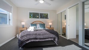 A bedroom with a bed, two bedside lamps, a ceiling fan, a window with a view, and a door leading to another room.