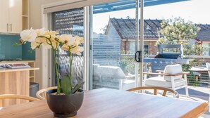 A modern kitchen with a wooden table, a potted orchid, and a sliding glass door leading to an outdoor patio with a grill. - Alkira by Experience Jervis Bay (Huskisson)