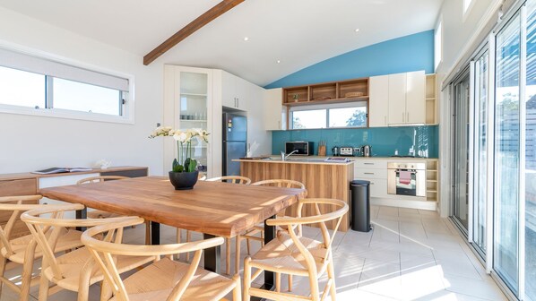 A modern kitchen with a wooden dining table and chairs, a built-in oven, and a large window providing natural light.
