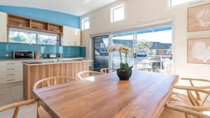 A modern kitchen with a wooden island, a dining area with a wooden table and chairs, and a view of the backyard through large sliding glass doors. - Alkira by Experience Jervis Bay (Huskisson)