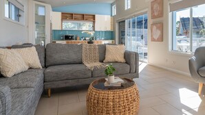 A modern living room with a grey sofa, a woven coffee table, and a view of the kitchen and outside. - Alkira by Experience Jervis Bay (Huskisson)