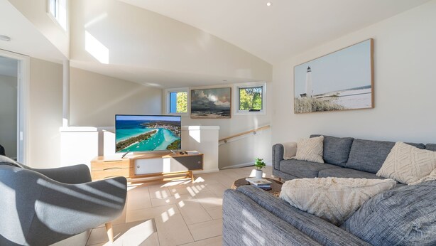 A modern living room with a large flat-screen TV, a grey sofa, and a coffee table. There are three framed pictures on the wall, and a staircase is visible in the background. - Alkira by Experience Jervis Bay (Huskisson)