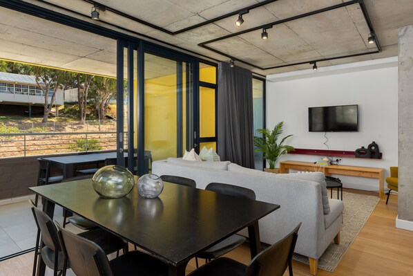 A modern living room with a dining area, large windows, a flat-screen TV, and a view of the outdoors.