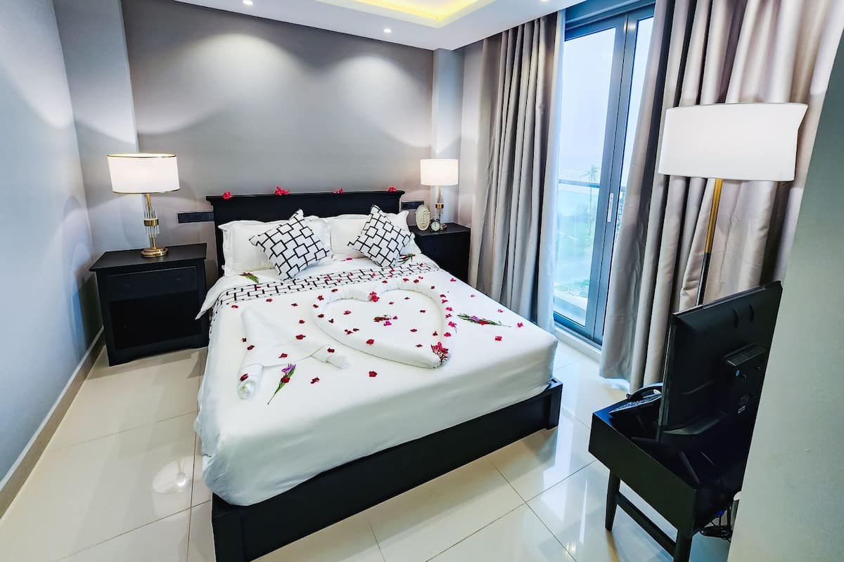 Una habitación de hotel moderna con una cama grande, dos mesitas de noche con lámparas, un ventilador de techo y vistas al exterior a través de un ventanal.