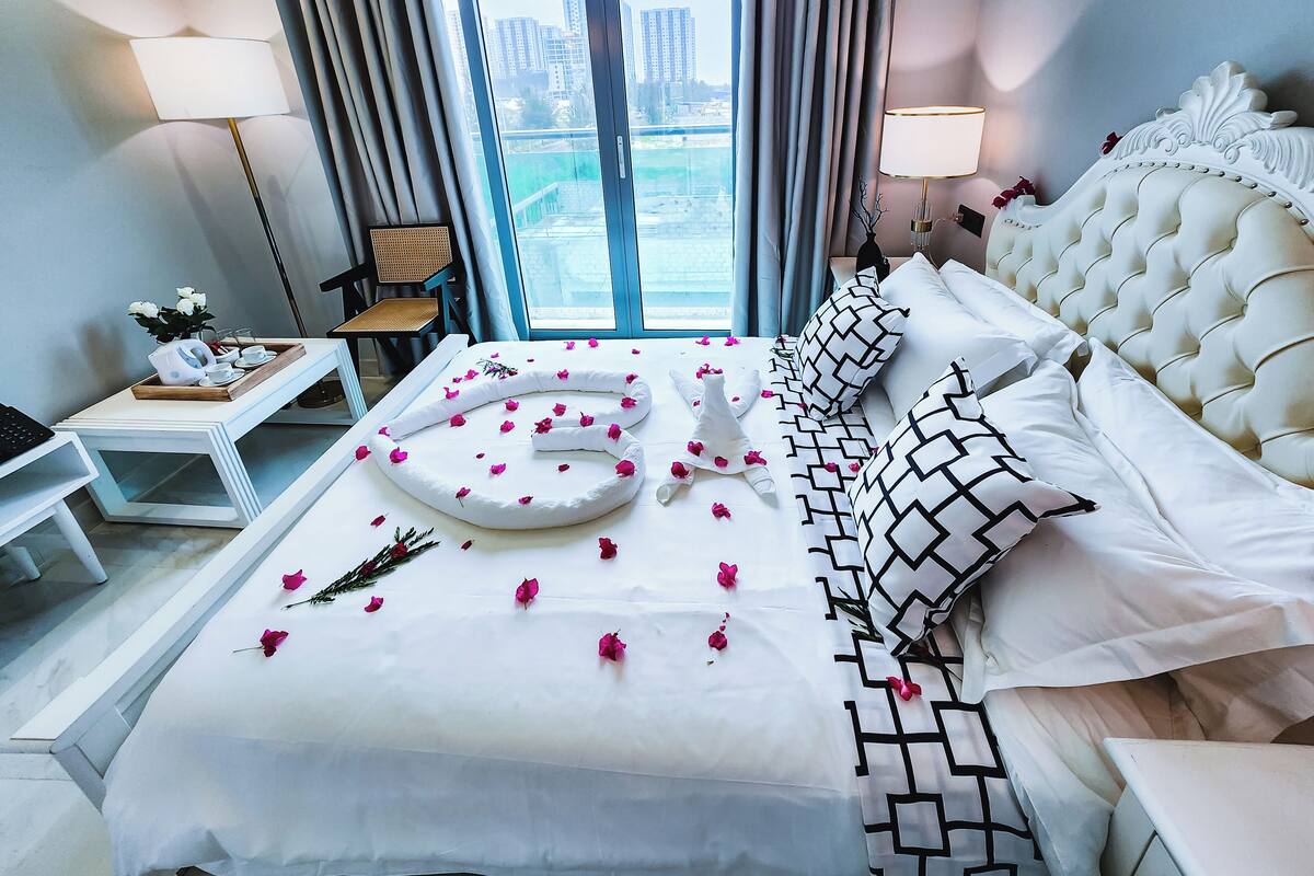 Habitación de hotel con cama, mesita de noche, una silla y vistas a la ciudad a través de amplias ventanas.