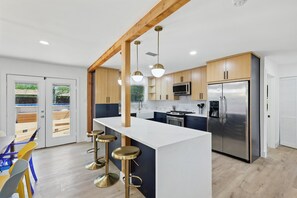 A modern kitchen with a central island, bar stools, wooden cabinets, stainless steel appliances, and a view of the outdoors through glass doors.