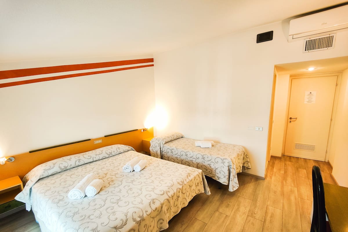A hotel room with two beds, each with a patterned bedspread and rolled towels. There is a wooden floor, a chair, and a door with a vent.