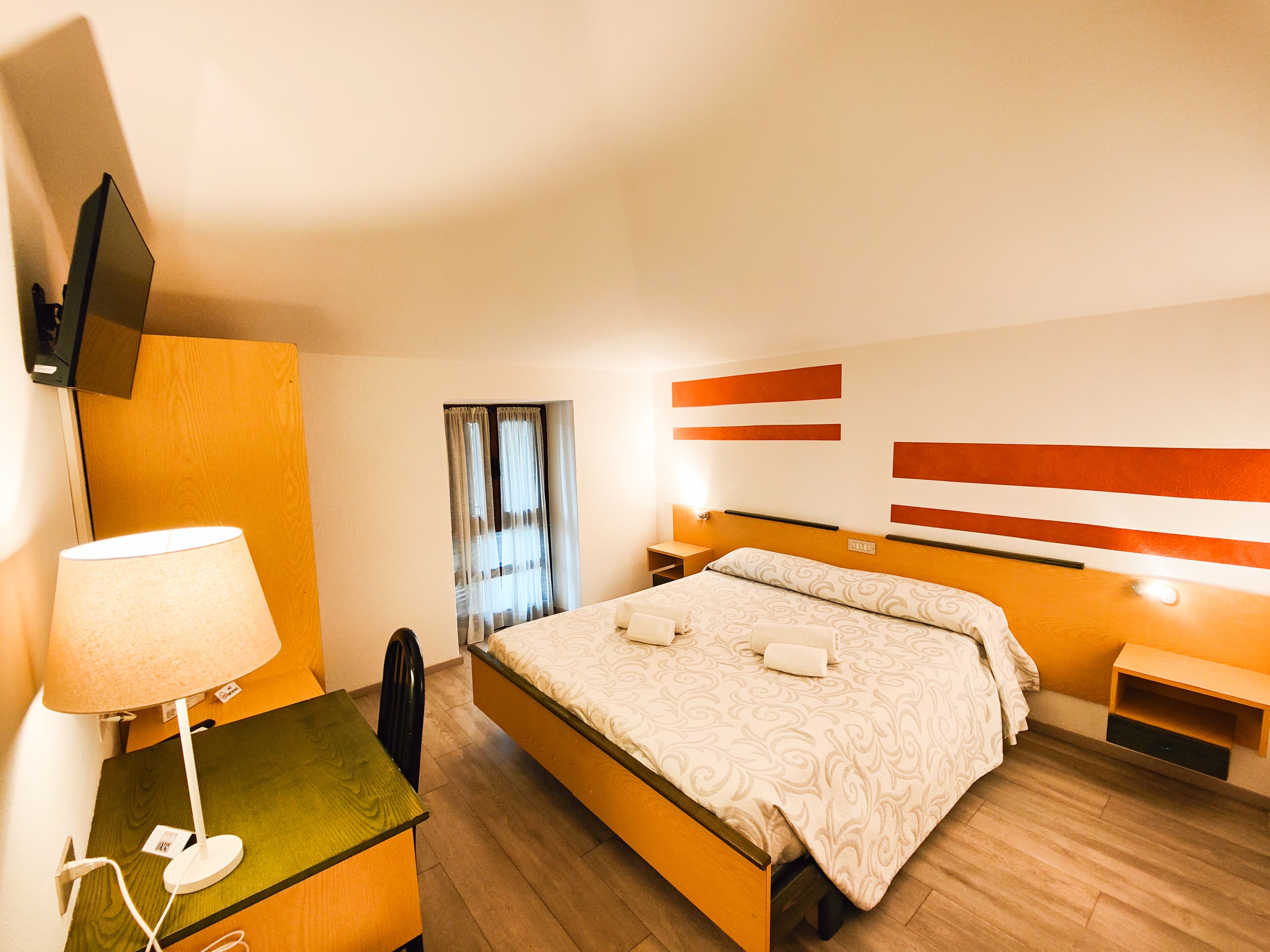 Camera d'albergo con un letto, comodini, una scrivania con lampada, una sedia, una TV a parete e una finestra con tende.