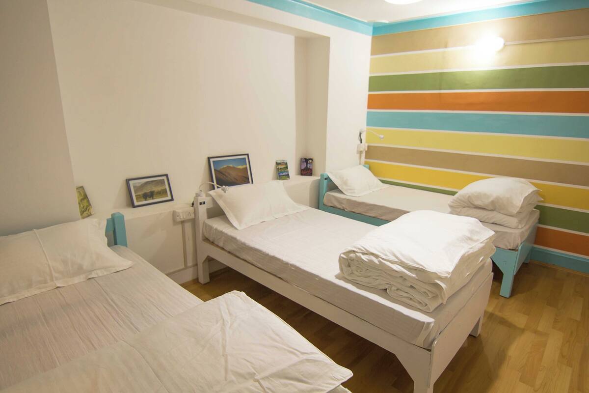 A room with two single beds, each with white bedding and pillows, a small bedside table, and a wall-mounted shelf with a framed picture and a small decorative item.