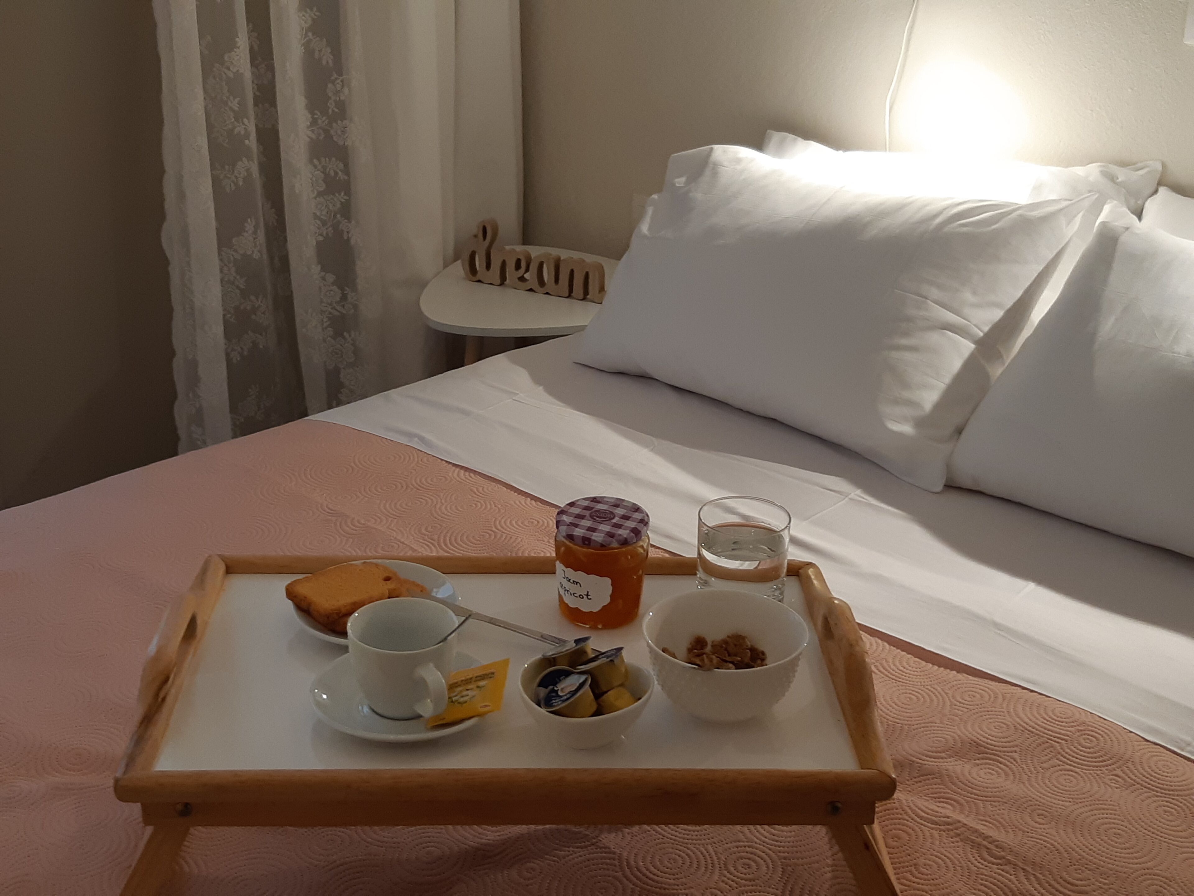 En pænt anrettet bakke med morgenmad på sengen, med et rosa sengetæppe, inklusiv en kop, et syltetøjsglas, et glas vand og en skål med müsli.