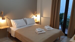 Ein ordentlich bezogenes Bett mit weißen Laken und zwei gefalteten Handtüchern, ein Nachttisch aus Holz mit einer Lampe und Blick ins Grüne durch eine Glastür.