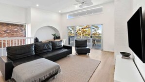 A modern living room with a black leather sofa, a flat-screen TV, and a balcony with glass doors. - Three Beaches (Malua Bay)
