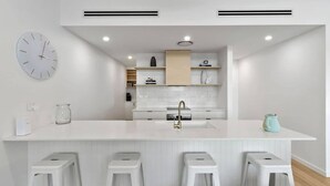 A modern kitchen with a central island, bar stools, a clock, and built-in shelves. - Three Beaches (Malua Bay)
